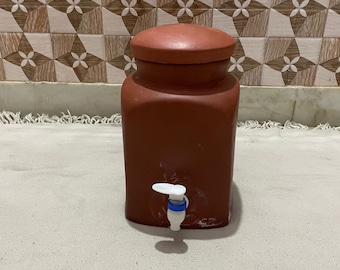 May include: A terracotta water dispenser with a white tap and a lid. The dispenser is sitting on a white surface.