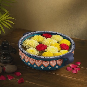 May include: A blue decorative bowl with a floral design, filled with water and yellow and red flowers. The bowl has two handles and is sitting on a wooden surface. Scattered rose petals are nearby.