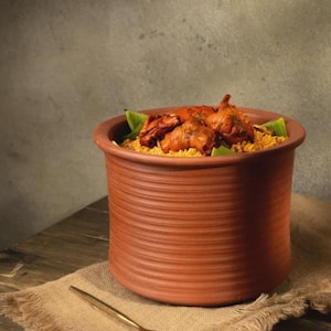 May include: A terracotta pot filled with seasoned rice and topped with cooked chicken and green leaves. A gold spoon rests on a burlap cloth next to the pot. A small plate with sliced tomatoes and onions is also visible.