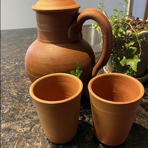 Könnte beinhalten: Ein Terrakotta-Krug mit Deckel und Henkel, zusammen mit zwei passenden Terrakotta-Tassen. Der Krug und die Tassen haben eine warme, erdige braune Farbe. Das Set ist wahrscheinlich handgefertigt und für Getränke geeignet.