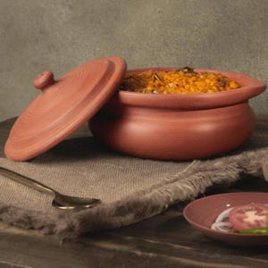May include: A terracotta clay pot with a lid, filled with a yellow-orange dish. A small plate with sliced tomatoes, onions, and a green chili is in the foreground. A spoon rests on a gray cloth.