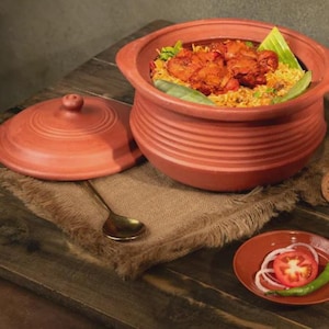 May include: A terracotta clay pot with a lid, filled with cooked food, sits on a wooden table. The pot has a round shape with horizontal ridges. A small plate with sliced tomatoes, onions, and a green chili is also on the table.