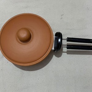 May include: A terracotta cooking pot with a lid and two black metal handles. The pot is round and has a small knob on the lid.