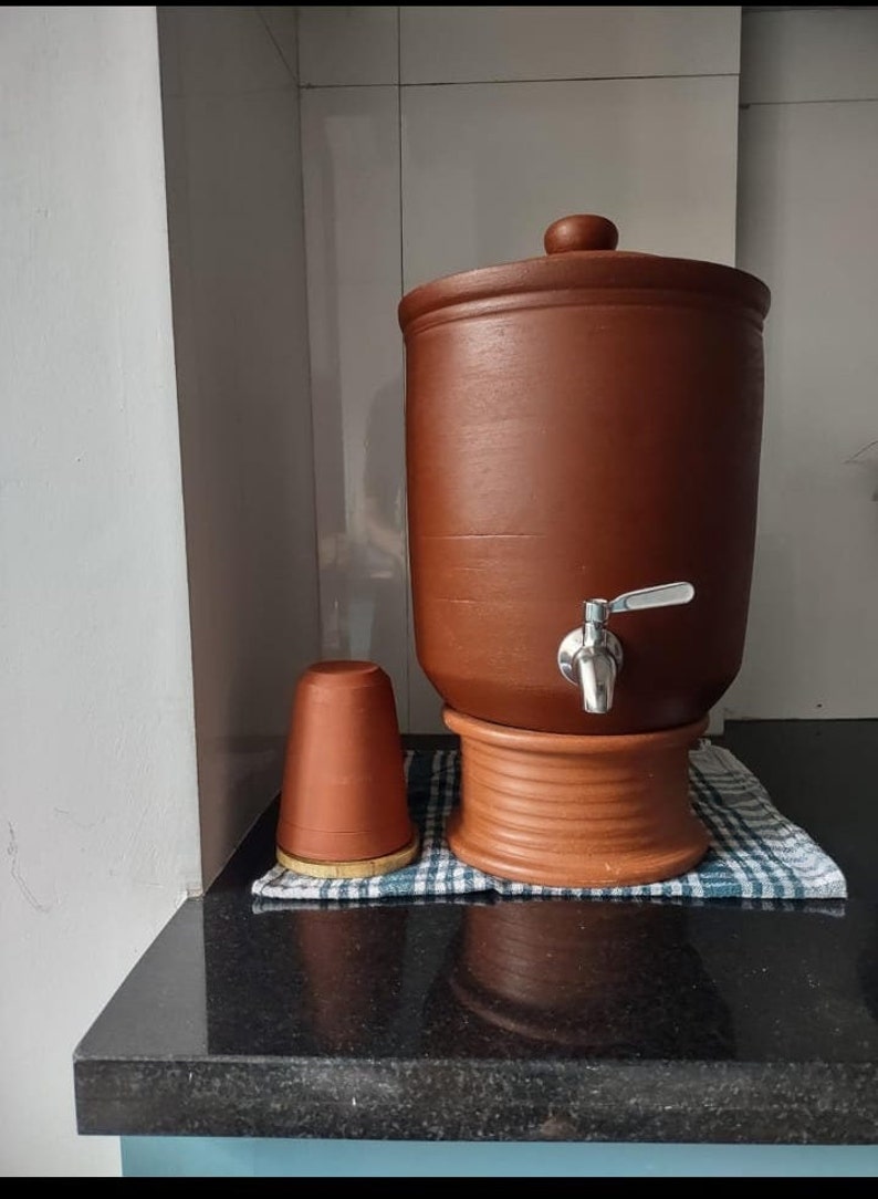 K&ouml;nnte beinhalten: Ein Terrakotta-Wasserfilter mit einem Hahn und einer passenden Terrakotta-Tasse. Der Filter steht auf einer schwarzen Arbeitsplatte.