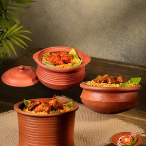 May include: Three terracotta clay pots filled with food, including one with a lid. The pots are arranged on a wooden surface with a neutral background. The food appears to be a rice dish with meat and garnishes.