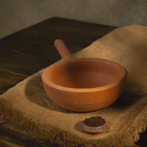 Può includere: Una ciotola color terracotta con un manico, appoggiata su un panno di iuta. Un piccolo contenitore rotondo con contenuto marrone scuro è vicino. Il design della ciotola suggerisce che potrebbe essere usata per servire o cucinare.