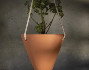 Puede incluir: Una maceta colgante en forma de cono de color terracota con una pequeña planta en su interior. La maceta está suspendida por una cuerda blanca. La planta tiene hojas verdes y blancas y pequeños capullos florales. El fondo es una pared gris texturizada.