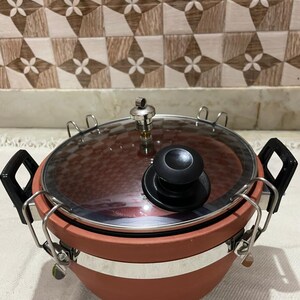 Könnte beinhalten: Ein Tonkochtopf mit einem Glasdeckel und einem Metallverschluss. Der Topf hat ein silbernes Band um die Oberseite und einen schwarzen Knopf auf dem Deckel.