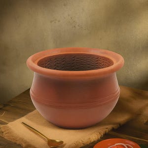 Puede incluir: Una olla de cocina de terracota marrón con un borde ancho y un cuerpo redondeado. Una pequeña cuchara dorada descansa sobre un paño beige junto a la olla. También hay un plato naranja pequeño con tomate en rodajas, cebolla y un chile verde.