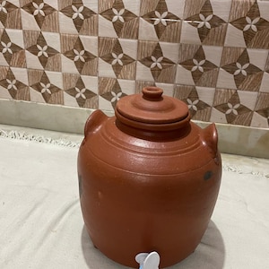 Puede incluir: Un dispensador de agua de terracota con un pico blanco. El dispensador tiene un cuerpo redondeado y una tapa con un pomo en la parte superior.