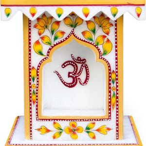 May include: A white marble altar with yellow and red accents. The altar features floral designs and the Om symbol. The design includes a shelf and a decorative top, showcasing intricate hand-painted details.