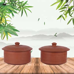 May include: Two terracotta clay pots with lids. The pots are a reddish-brown color and have a round shape with a lid and a small knob on top. The pots are sitting on a wooden surface with a background of green leaves and a mountain landscape.