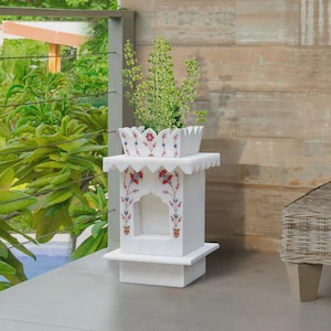Pode incluir: Um vaso de mármore branco com um design floral decorativo em vermelho, azul e verde. O vaso tem uma base quadrada e um topo detalhado, segurando uma planta verde. O design lembra a arquitetura tradicional indiana.