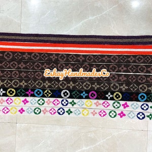 May include: A collection of 10 colourful fabric straps with different patterns, including a black and white monogram pattern, a colourful circle and flower pattern, and a solid red and white stripe pattern. The straps have gold metal clasps on each end.