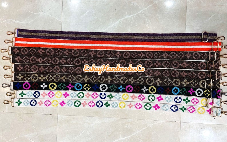 May include: A collection of 10 colourful fabric straps with different patterns, including a black and white monogram pattern, a colourful circle and flower pattern, and a solid red and white stripe pattern. The straps have gold metal clasps on each end.