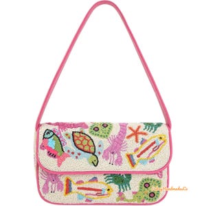 Puede incluir: Bolso de mano rectangular de cuentas de color blanco roto con correa y ribete rosas. El bolso presenta criaturas marinas de cuentas de colores, como peces, una tortuga y una langosta. El diseño está delineado en rosa.
