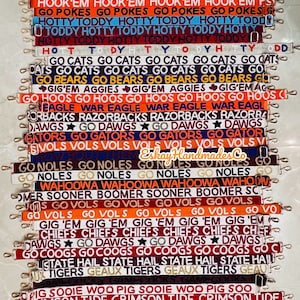 May include: A collection of 20 colorful woven straps with various college team logos and slogans. The straps feature a variety of colors, including red, blue, white, orange, black, and gold. The straps are all approximately 1 inch wide and have a gold metal clasp.