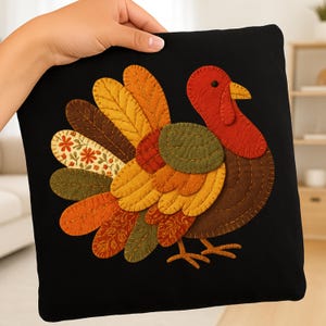 May include: Black square pillow featuring a colorful turkey design. The turkey has a red head, a brown body, and a vibrant tail with orange, yellow, and brown feathers. The pillow is held by a hand.