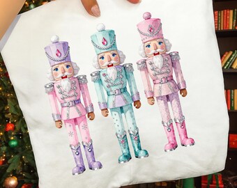 May include: A white pillowcase featuring a watercolor illustration of three toy nutcrackers. Each nutcracker is in a different pastel color: pink, blue, and purple. They have silver accents and are wearing tall hats.