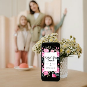 Op de afbeelding: Een smartphone die een uitnodiging met bloemenmotief voor een Moederdagbrunch laat zien. De uitnodiging bevat de tekst "Mother's Day Brunch" en details over het evenement. De telefoon staat op een tafel met een boeket witte bloemen in een glazen vaas.