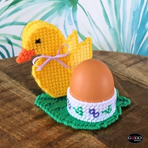 Pode incluir: Um patinho amarelo feito à mão com bico laranja e laço lilás, ao lado de um ovo castanho num suporte para ovo branco e azul, tudo sobre uma base verde em forma de folha. Ideal para a Páscoa.