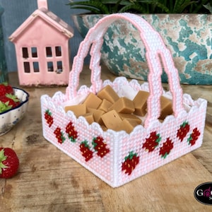 Op de afbeelding: Een roze plastic mandje met een witte handvat en een aardbeidesontwerp. Het mandje is gevuld met kleine vierkantjes karamel.