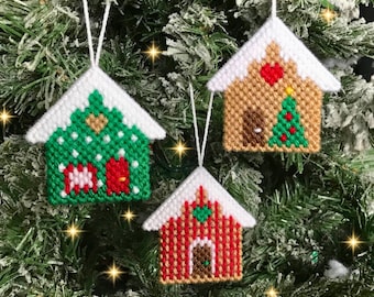 May include: Three handmade Christmas ornaments shaped like houses. The houses are decorated with red, green, and white yarn and have a snowy white roof. The ornaments are hanging on a green Christmas tree.
