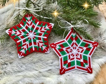 May include: Two red, green, and white star-shaped ornaments with a snowflake design. The ornaments are made of plastic canvas and have white ribbon loops for hanging.
