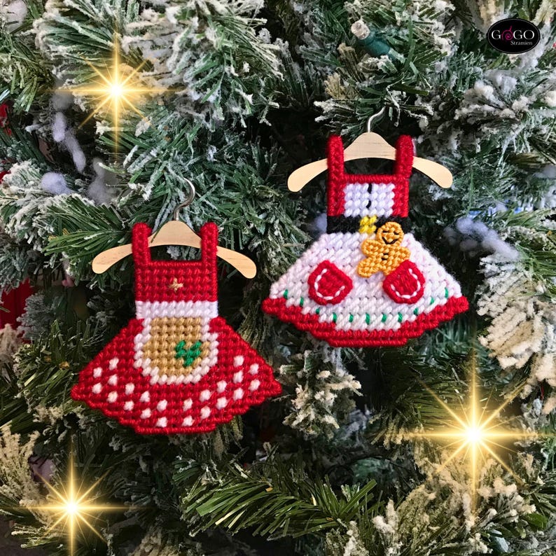 May include: Two red and white Christmas ornaments shaped like dresses with a gingerbread person design. The ornaments are hanging on a green Christmas tree.