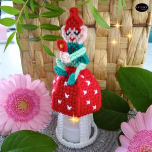 May include: A handmade crochet gnome candle holder. The gnome wears a red hat and a red mushroom-shaped skirt with white accents. The candle holder is white and has a lit candle inside. Pink flowers and greenery surround the gnome.