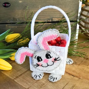 May include: A white plastic canvas Easter basket shaped like a bunny, with a handle and filled with red candies. The bunny has gray and pink accents, with a smiling face and black whiskers. The basket is sitting on a wooden surface.