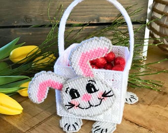 May include: A white plastic canvas Easter basket shaped like a bunny, with a handle and filled with red candies. The bunny has gray and pink accents, with a smiling face and black whiskers. The basket is sitting on a wooden surface.