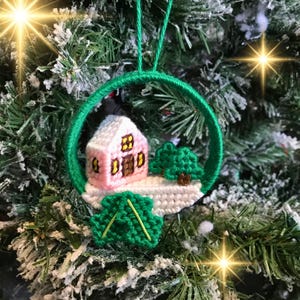 May include: A pink and white crocheted house ornament with a green tree and a green leaf, hanging from a green string on a Christmas tree.