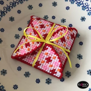 May include: A square, handmade needlepoint item in red, pink, and white floral pattern, tied with a yellow ribbon. The item is on a white plate with blue floral designs. The needlepoint has a red border.