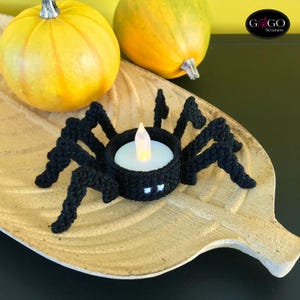 May include: A black crocheted spider tea light holder with a white LED candle, set on a leaf-shaped tray. Two yellow pumpkins are in the background. The tea light holder has white eyes and is a Halloween decoration.