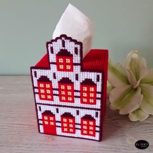 Puede incluir: Un cubretisú decorativo con forma de casa, con tejado y paredes rojas y detalles blancos. Las ventanas son rojas con detalles amarillos. Un pañuelo blanco es visible en la parte superior. Diseño de punto de cruz.