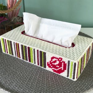 Puede incluir: Cubierta de caja de pañuelos blanca con un diseño de flor roja y un borde de rayas de colores. La cubierta está hecha de hilo y tiene una ranura para pañuelos.