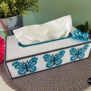 May include: A white tissue box cover with a blue and gray butterfly design. The cover is made of plastic canvas and has a slot for tissues. The box is sitting on a gray woven surface.