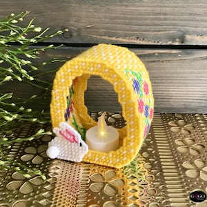 May include: A yellow and white crocheted Easter egg shaped candle holder with a white bunny peeking out from the bottom. The candle holder has a floral design and is lit with a small white tea light.
