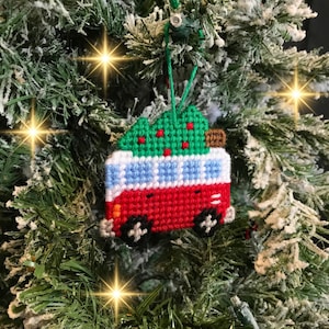May include: A handmade Christmas ornament shaped like a red, white, and blue camper van with a green Christmas tree on top. The ornament is made of yarn and has black wheels. It hangs from a green string.
