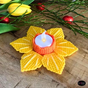 以下が含まれることがあります： 手作りの、黄色い編み物の花型のキャンドルホルダー。オレンジ色の中心部には白いLEDキャンドルが入っています。背景には黄色のチューリップ、緑の小枝、赤い包み紙のキャンディーがあります。