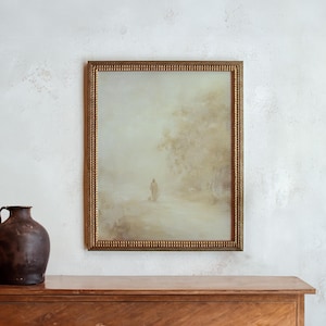 May include: A framed landscape painting with a gold-colored ornate frame. The artwork depicts a figure walking on a path in a misty, muted-color scene. A dark brown jug sits on a wooden surface to the left of the painting.