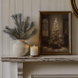 May include: A white ceramic vase with a green pine sprig arrangement sits on a white mantel with a framed picture of a Christmas tree and a white candle on a gold metal plate.