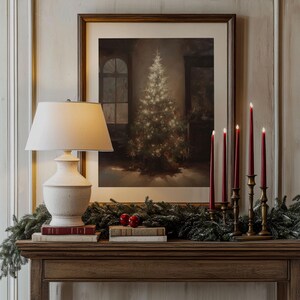 May include: A white lamp with a cream-colored shade sits on a wooden table with a framed print of a Christmas tree.  The table is decorated with a garland of evergreen branches and red ornaments.  Four gold candle holders with red candles are on the table.