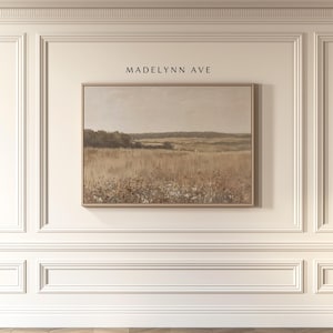 May include: A framed landscape painting with a neutral color palette. The artwork depicts a field of tall grass and wildflowers under a muted sky. The text "MADELYNN AVE" is above the painting. The frame is a light wood color.