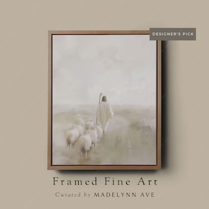 May include: A framed painting of a man in white robes walking with a flock of sheep in a field. The sky is a soft white with hints of blue. The painting is framed in a light brown wood frame.