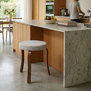 Könnte beinhalten: Eine moderne Küche mit einer Kücheninsel aus Marmor und Holzschränken. Ein grauer Hocker mit Holzbeinen steht vor der Insel. Ein Esstisch mit Stühlen ist durch ein großes Fenster zu sehen. Eine Person ist im Hintergrund.