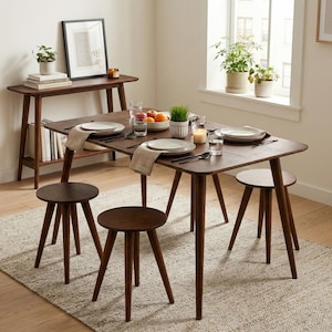 May include: A dark wood dining table with four matching round stools, featuring tapered legs. The table is set with white plates, linen napkins, glassware, a bowl of fruit, a small potted plant, and a candle. A matching console table stands nearby.