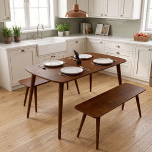 May include: A dark brown wooden dining table with rounded corners and two matching benches. The table is set with four white plates, four sets of silverware, and a small dark vase in the center. The furniture features tapered legs.