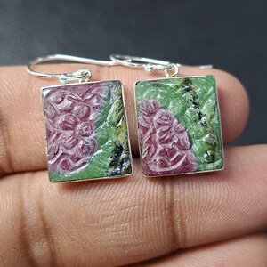 May include: A pair of rectangular earrings with a floral design carved into the ruby zoisite stones. The earrings have a silver-colored metal border and ear hooks. The stones feature a mix of pink, green, and black colors.